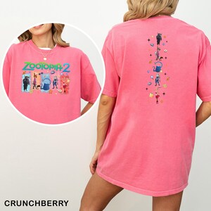 May include: A pink t-shirt featuring the text "ZOOTOPIA 2" with cartoon characters. The back of the shirt has a vertical design of cartoon characters. The shirt is a solid pink color and has short sleeves.