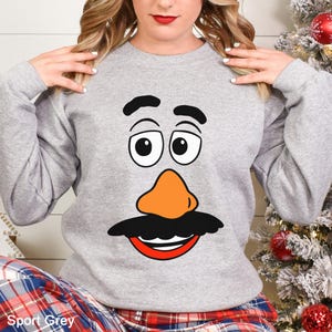 May include: Sport Grey sweatshirt featuring a cartoon face with black eyebrows, eyes, mustache, and an orange nose. The design is centered on the chest. The sweatshirt has long sleeves and a crew neck. The text "Sport Grey" is at the bottom.