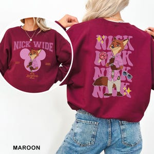 K&ouml;nnte beinhalten: Burgunderfarbenes Sweatshirt mit einem Cartoon-Fuchs-Design. Auf der Vorderseite stehen "NICK WIDE" und "Zootopia" mit einer Fuchsgrafik. Auf der R&uuml;ckseite befindet sich eine gr&ouml;&szlig;ere Fuchsgrafik und der Name "Nick" wiederholt.