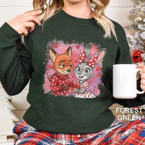 K&ouml;nnte beinhalten: Dunkelgr&uuml;nes Sweatshirt mit einem Cartoon-Fuchs und Hasen, die ein herzf&ouml;rmiges Geschenk halten. Der Fuchs tr&auml;gt einen roten Schal und der Hase eine rote Schleife. Rote Herzen auf rosa Hintergrund.