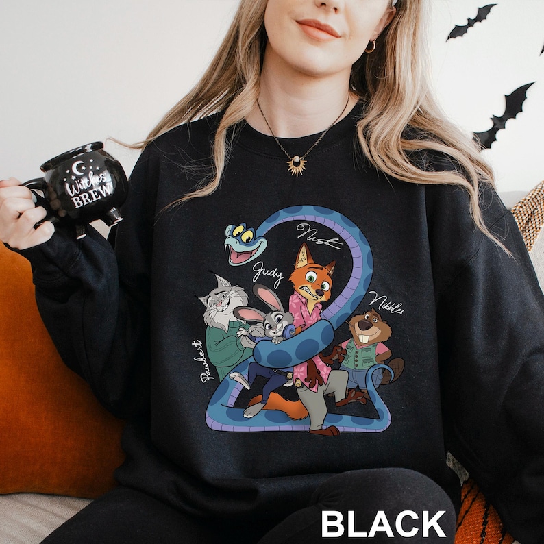 Op de afbeelding: Zwarte sweatshirt met een cartoon illustratie van dierenfiguren, waaronder een vos, een konijn en een bever, met de namen "Judy", "Nick" en "Nibbles". Het woord "BLACK" staat onderaan gedrukt.