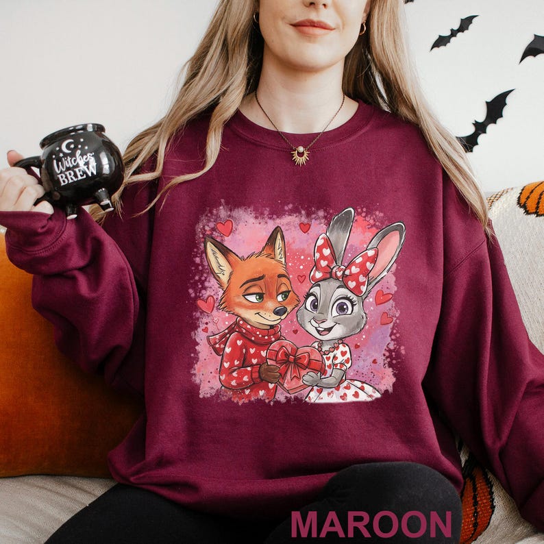 K&ouml;nnte beinhalten: Burgunderfarbenes Sweatshirt mit einem Cartoon-Fuchs und -Hasen, die ein herzf&ouml;rmiges Geschenk halten. Der Fuchs tr&auml;gt einen roten Pullover mit wei&szlig;en Herzen, und der Hase hat eine rote Schleife. Das Wort "MAROON" ist unten aufgedruckt.