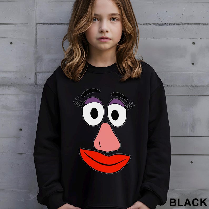 May include: Black sweatshirt featuring a cartoon face with large white eyes, purple eyeshadow, a pink nose, and a red mouth. The word "BLACK" is printed in white at the bottom right corner. The sweatshirt is worn by a person.