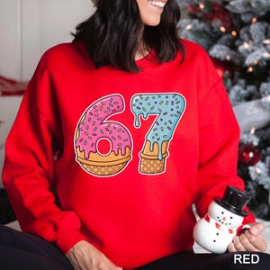 May include: A red sweatshirt featuring a graphic of the number "67" designed as a donut and ice cream cone. The "6" is a pink donut with sprinkles, and the "7" is blue ice cream with sprinkles. The word "RED" is in the bottom right corner.
