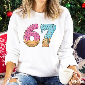 May include: White sweatshirt featuring a colorful graphic of the number "67" designed as a donut and ice cream cone. The "6" is pink with sprinkles, and the "7" is blue with sprinkles. The sweatshirt is a casual, comfortable style.