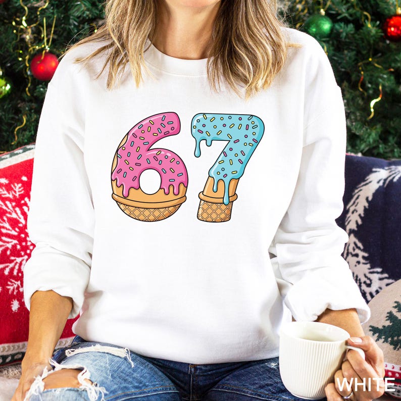 May include: White sweatshirt featuring a colorful graphic of the number "67" designed as a donut and ice cream cone. The "6" is pink with sprinkles, and the "7" is blue with sprinkles. The sweatshirt is a casual, comfortable style.
