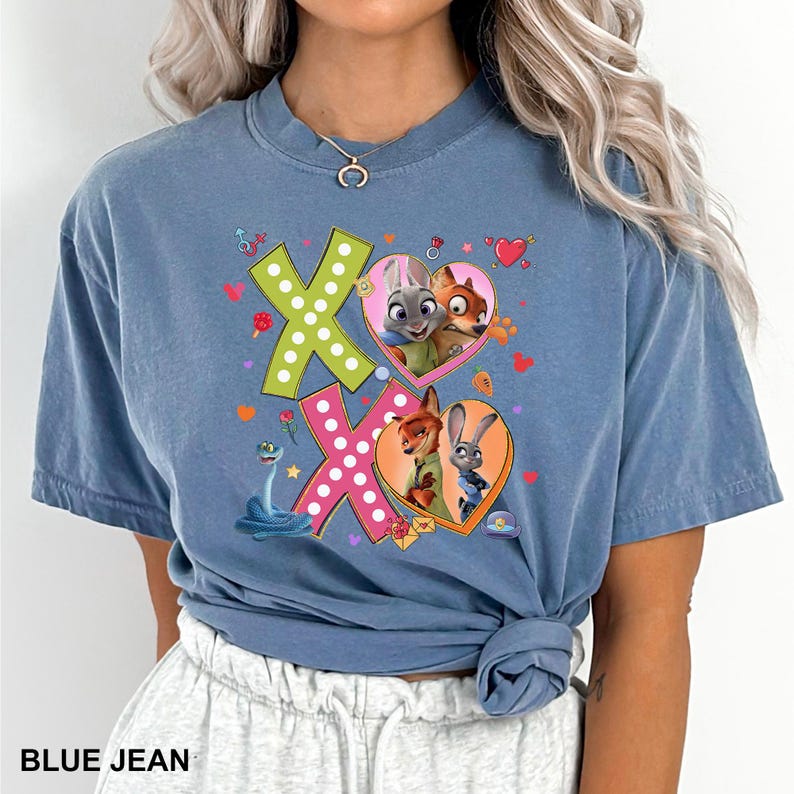 May include: Blue jean colored t-shirt featuring a graphic design with the text "XO". The design includes cartoon characters in heart-shaped frames, a snake, and other decorative elements. The shirt is a casual style.