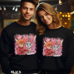 K&ouml;nnte beinhalten: Schwarze Sweatshirts mit einem Cartoon-Fuchs- und Hasen-Design. Der Fuchs tr&auml;gt einen roten Schal und der Hase hat eine Schleife mit Herzen. Sie halten ein herzf&ouml;rmiges Geschenk. Das Design ist vor einem rosa Hintergrund mit Herzen.