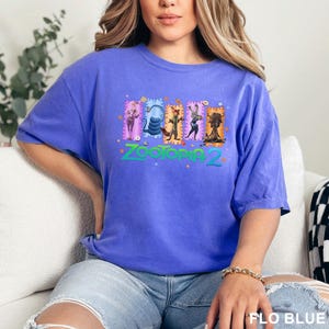 K&ouml;nnte beinhalten: Ein kurz&auml;rmeliges T-Shirt in Kornblumenblau mit einem Grafikdruck von f&uuml;nf animierten Charakteren aus Zootopia 2, die in rechteckigen Rahmen &uuml;ber dem t&uuml;rkisfarbenen Schriftzug Zootopia 2 angeordnet sind, akzentuiert durch kleine orangefarbene Pfotenabdr&uuml;cke.