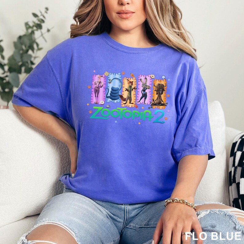 K&ouml;nnte beinhalten: Ein kurz&auml;rmeliges T-Shirt in Kornblumenblau mit einem Grafikdruck von f&uuml;nf animierten Charakteren aus Zootopia 2, die in rechteckigen Rahmen &uuml;ber dem t&uuml;rkisfarbenen Schriftzug Zootopia 2 angeordnet sind, akzentuiert durch kleine orangefarbene Pfotenabdr&uuml;cke.