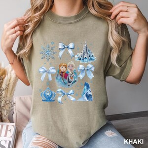 May include: Khaki-colored t-shirt featuring a design with characters and elements from the animated film. The design includes images of Anna, Elsa, Olaf, snowflakes, bows, a castle, and an ice formation. The word "KHAKI" is printed at the bottom right.