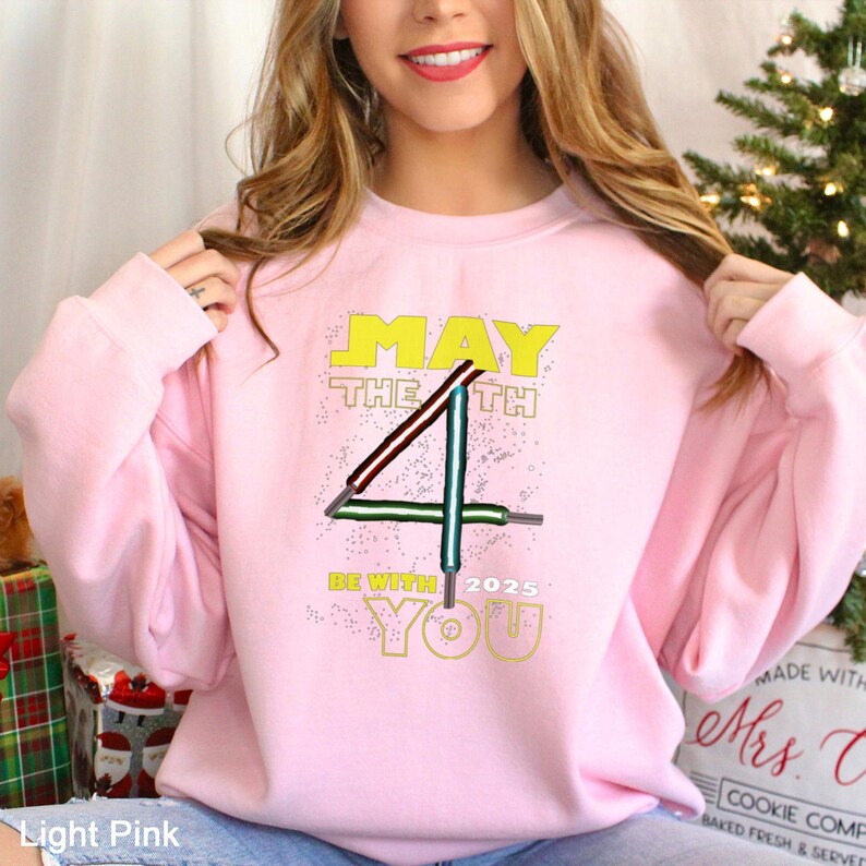 May include: Pale pink sweatshirt featuring the phrase "MAY THE 4TH BE WITH YOU 2025" in yellow, with a graphic of lightsabers forming the number 4. The sweatshirt has a crew neck.