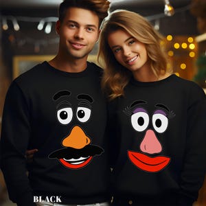 May include: Black sweatshirts featuring Mr. and Mrs. Potato Head faces. The Mr. Potato Head sweatshirt has a black mustache, orange nose, and red mouth. The Mrs. Potato Head sweatshirt has purple eyelashes, a pink nose, and a red mouth.