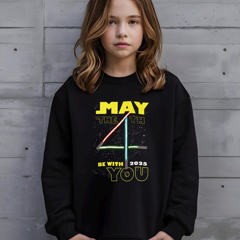 May include: Black sweatshirt featuring the text "MAY THE 4TH BE WITH YOU 2025" in yellow and white. The design includes a stylized number 4 with red and green lightsaber-like lines. The sweatshirt is worn by a person.