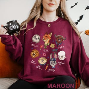 May include: Maroon sweatshirt featuring cartoon characters from Zootopia. The design includes Flash, Nick Wilde, Chief Bogo, Mayor Lionheart, Officer Clawhauser, Judy Hopps, and Assistant Bellwether. The word "MAROON" is printed at the bottom of the sweatshirt.