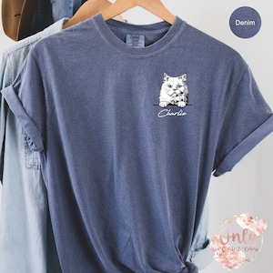 May include: A denim blue t-shirt with a white cat peeking out of a pocket and the name "Charlie" written below.