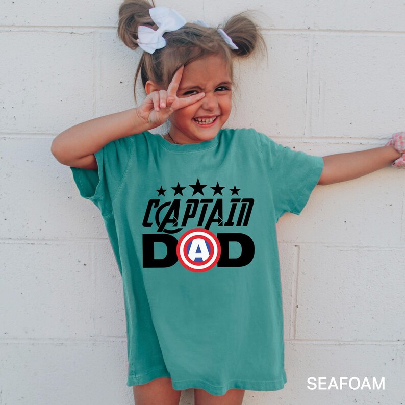 Puede incluir: Camiseta de color verde azulado con la frase "CAPTAIN DAD" en blanco y negro, con un emblema de escudo rojo y blanco. El dise&ntilde;o incluye cinco estrellas negras encima del texto. La palabra "SEAFOAM" est&aacute; impresa en la esquina inferior derecha.