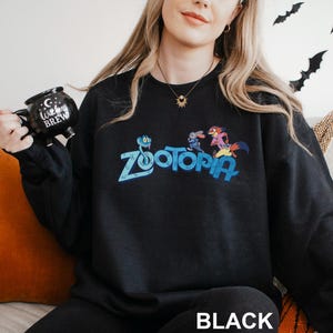 Puede incluir: Sudadera negra de Zootopia con el logotipo de Zootopia y personajes de dibujos animados. La sudadera tiene cuello redondo y mangas largas. La palabra "BLACK" est&aacute; impresa en la parte inferior de la imagen.
