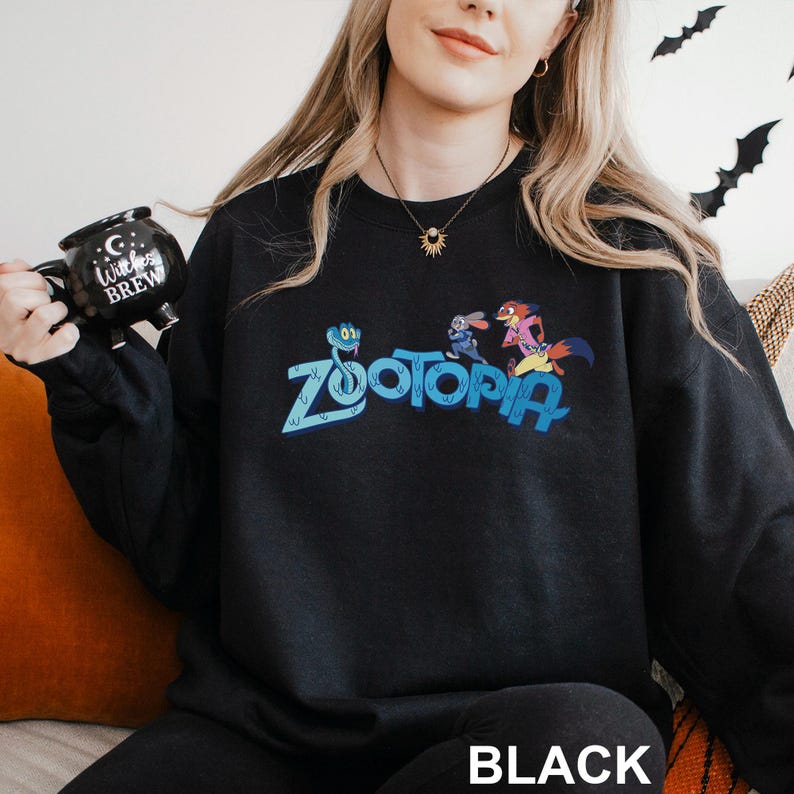 Puede incluir: Sudadera negra de Zootopia con el logotipo de Zootopia y personajes de dibujos animados. La sudadera tiene cuello redondo y mangas largas. La palabra "BLACK" est&aacute; impresa en la parte inferior de la imagen.