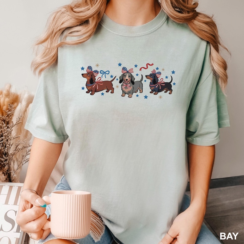 Peut inclure: T-shirt vert clair avec trois teckels portant des n&oelig;uds patriotiques et des &eacute;toiles bleues. Les teckels sont marron, gris et noir. Le t-shirt est de style d&eacute;contract&eacute;.