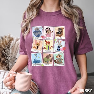 May include: Berry-colored t-shirt featuring a grid of cartoon character portraits. Each portrait is in a polaroid-style frame with character names like Chief Bogo, Judy, and Mr. Big. The shirt's color is labeled "BERRY" at the bottom.