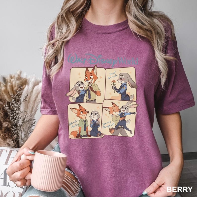 K&ouml;nnte beinhalten: Ein beerenfarbenes Kurzarm-T-Shirt mit einem Walt Disney World-Grafikdruck, der in vier Feldern die Zeichentrickfiguren Nick Wilde und Judy Hopps in verschiedenen Posen zeigt, inklusive der Schriftz&uuml;ge Sweet Surrender, Adventure Awaits und Dancing Through Life.