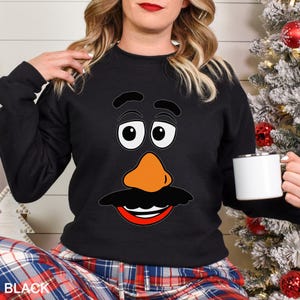 May include: Black sweatshirt featuring a cartoon face with large white eyes, an orange nose, and a black mustache and mouth. The word "BLACK" is visible at the bottom left. A white mug is held in the right hand.