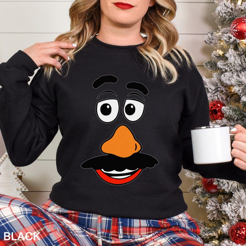 May include: Black sweatshirt featuring a cartoon face with large white eyes, an orange nose, and a black mustache and mouth. The word "BLACK" is visible at the bottom left. A white mug is held in the right hand.