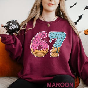 May include: Maroon sweatshirt featuring a colorful graphic of the number 67 designed as a donut and ice cream cone. The number 6 is a pink donut with sprinkles, and the 7 is blue ice cream with sprinkles. The word "MAROON" is at the bottom.