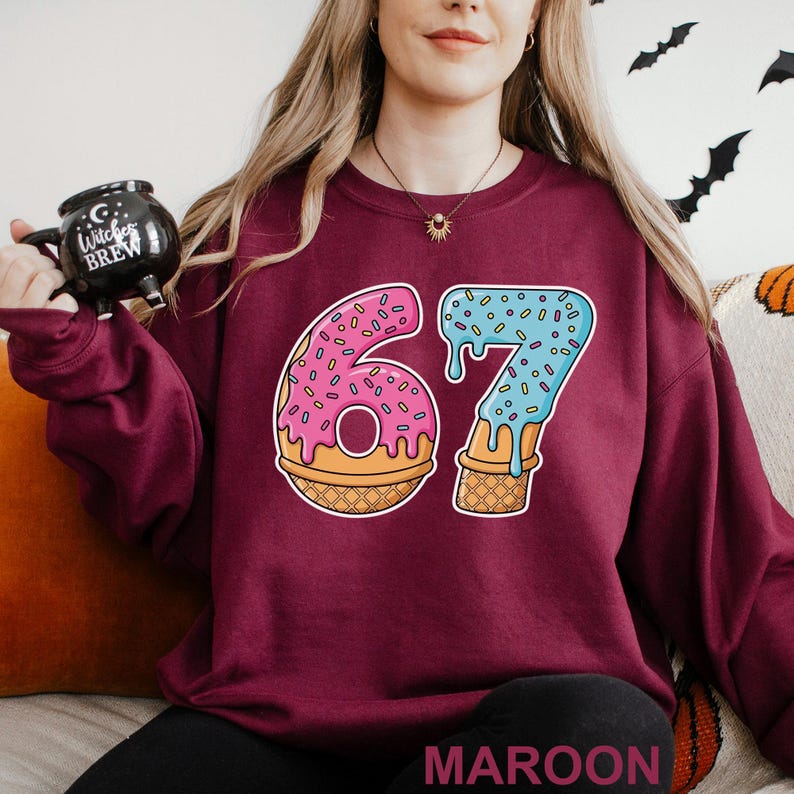 May include: Maroon sweatshirt featuring a colorful graphic of the number 67 designed as a donut and ice cream cone. The number 6 is a pink donut with sprinkles, and the 7 is blue ice cream with sprinkles. The word "MAROON" is at the bottom.