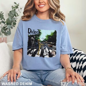 May include: Light blue t-shirt featuring a graphic of Star Wars droids crossing a street, with the text "The Droids Imperial Road". The shirt is made of a soft material and has the words "Washed Denim" printed at the bottom.