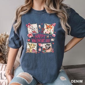 May include: Denim blue t-shirt featuring four cartoon character portraits with Valentine's Day themed elements. The words "HAPPY VALENTINE'S DAY" are printed in pink. The shirt is made of a soft, comfortable material.