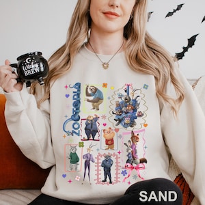 Puede incluir: Sudadera de color arena con un gr&aacute;fico colorido de personajes de la pel&iacute;cula animada "Zootopia". El dise&ntilde;o incluye varios personajes de animales en cuadros enmarcados, con la palabra "Zootopia" en una fuente estilizada. La palabra "SAND" est&aacute; impresa en la parte inferior.