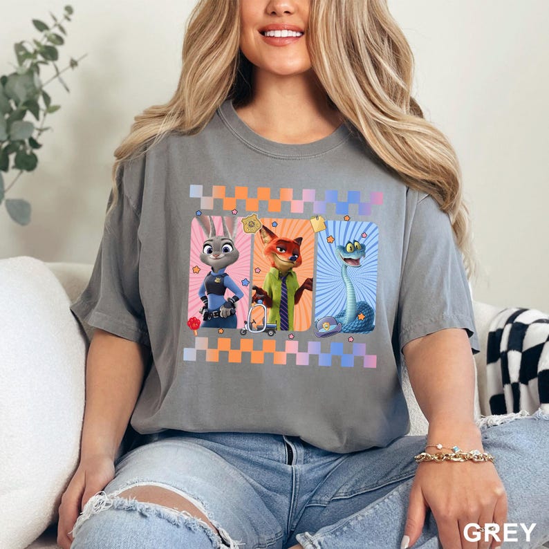May include: A charcoal gray short-sleeve t-shirt featuring a graphic print of three animated characters from Zootopia. The design includes Judy Hopps, Nick Wilde, and a blue snake, framed by colorful checkered borders and small star accents.