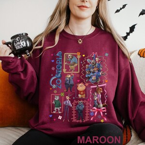 Puede incluir: Una sudadera granate con un gr&aacute;fico colorido de personajes de Zootopia. El dise&ntilde;o incluye im&aacute;genes enmarcadas de animales, la palabra "Zootopia" y la palabra "MAROON" en la parte inferior. La persona sostiene una taza negra.