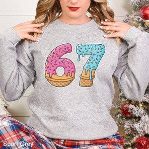 May include: A light gray sweatshirt with the number "67" in a playful, cartoon style. The "6" is designed as a pink donut with sprinkles, and the "7" is blue with sprinkles, resembling melting ice cream. The sweatshirt is labeled "Sport Grey".