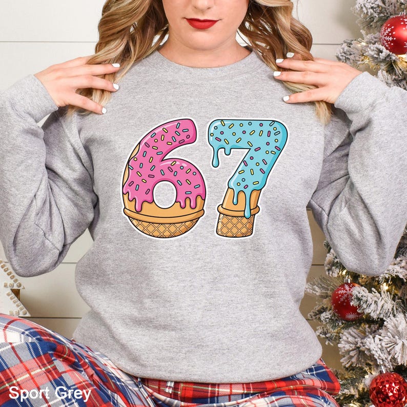 May include: A light gray sweatshirt with the number "67" in a playful, cartoon style. The "6" is designed as a pink donut with sprinkles, and the "7" is blue with sprinkles, resembling melting ice cream. The sweatshirt is labeled "Sport Grey".