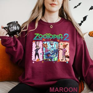 Puede incluir: Sudadera burdeos con cuello redondo con el texto "ZOOTOPIA 2" sobre un gr&aacute;fico de personajes de la pel&iacute;cula. El gr&aacute;fico incluye un conejo, un zorro y otros personajes. La palabra "MAROON" est&aacute; impresa en la parte inferior.