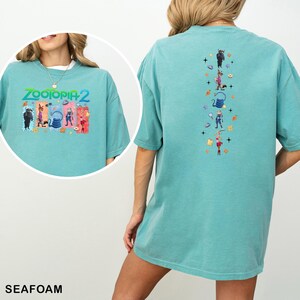 May include: Seafoam green t-shirt featuring the text "ZOOTOPIA 2" with cartoon characters on the front. The back of the shirt has a vertical design with more cartoon characters and symbols. The shirt is made of a soft material.