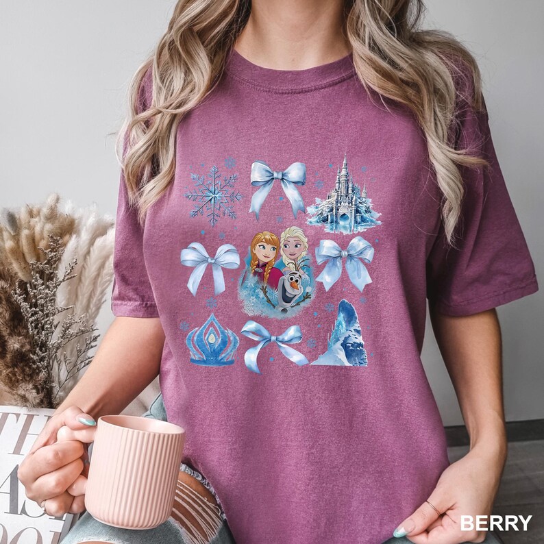 May include: Berry-colored t-shirt featuring a graphic design with images of a castle, snowflakes, bows, and characters from a popular animated movie. The word "BERRY" is printed in the bottom right corner of the shirt.