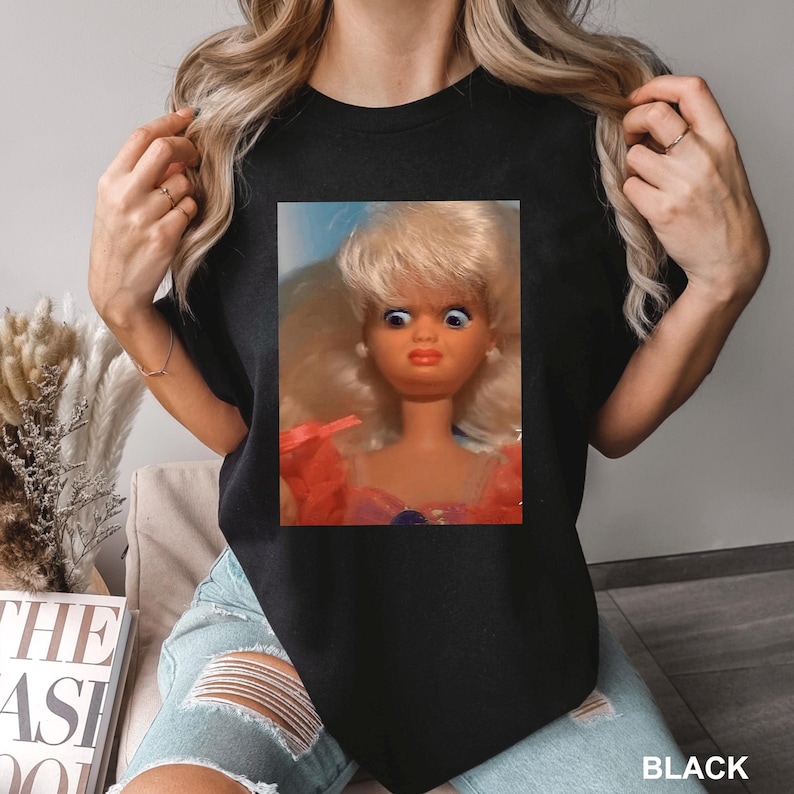 May include: Black t-shirt featuring a framed image of a vintage doll with a surprised expression. The doll has blonde hair, blue eyes, and is wearing a pink dress. The word "BLACK" is printed at the bottom right.
