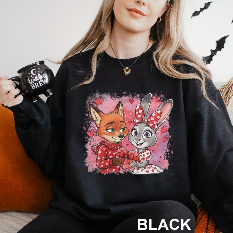 K&ouml;nnte beinhalten: Schwarzes Sweatshirt mit einem Cartoon-Fuchs und -Hasen, die ein herzf&ouml;rmiges Geschenk teilen. Der Fuchs tr&auml;gt einen roten Schal, der Hase hat eine rote Schleife. Das Wort "BLACK" ist unten aufgedruckt.