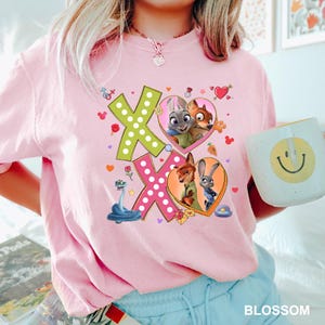 May include: A light pink t-shirt featuring the characters from Zootopia. The shirt has a graphic design with the letters "XO" in green and pink, with images of the characters inside hearts. A mug with a smiley face is held in the image.