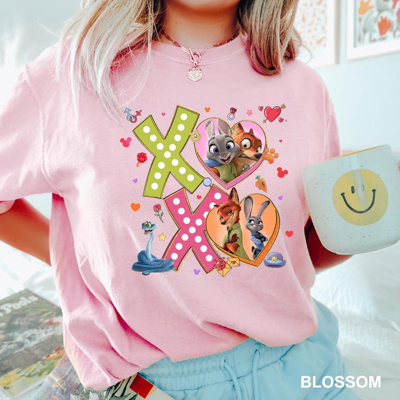 May include: A light pink t-shirt featuring the characters from Zootopia. The shirt has a graphic design with the letters "XO" in green and pink, with images of the characters inside hearts. A mug with a smiley face is held in the image.