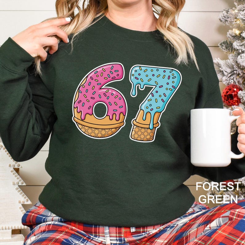 May include: Forest green sweatshirt featuring the number "67" designed as donuts and ice cream cones. The "6" is pink with sprinkles, and the "7" is blue with sprinkles. The sweatshirt is a solid color and has a crew neck.