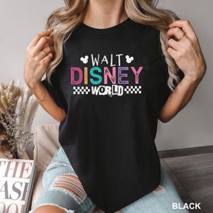 May include: A black short-sleeved t-shirt featuring the text WALT DISNEY WORLD in a multi-colored, distressed font. The design includes small white Mickey Mouse head silhouettes above the text and a black and white checkered pattern below.