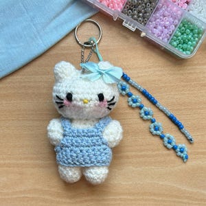 Peut inclure: Porte-clés Hello Kitty en crochet blanc avec une robe en jean bleu et un nœud bleu clair. Le porte-clés est attaché à une chaîne argentée avec des fleurs en perles bleu clair et bleu foncé.