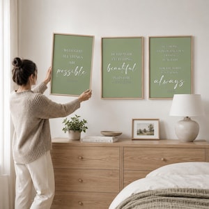 May include: Three framed art prints with inspirational quotes. The prints have a sage green background and wooden frames. The quotes are in white script. A woman is holding one of the prints. A lamp and a small framed picture are on a wooden dresser.