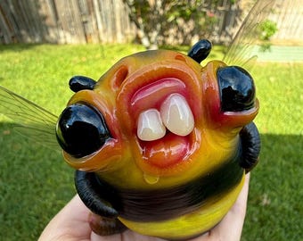 Peut inclure: Une sculpture en verre représentant une abeille au corps rond rayé jaune et noir, dotée de grands yeux noirs et de deux dents blanches proéminentes dans une bouche ouverte. La figurine possède des ailes translucides et des antennes.