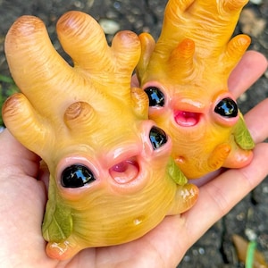 May include: Two quirky, yellow-orange sculptures with large, black eyes and smiling pink mouths. The sculptures have a unique, organic shape with multiple finger-like protrusions. Green leaf-like details are visible. The sculptures are held in a hand.